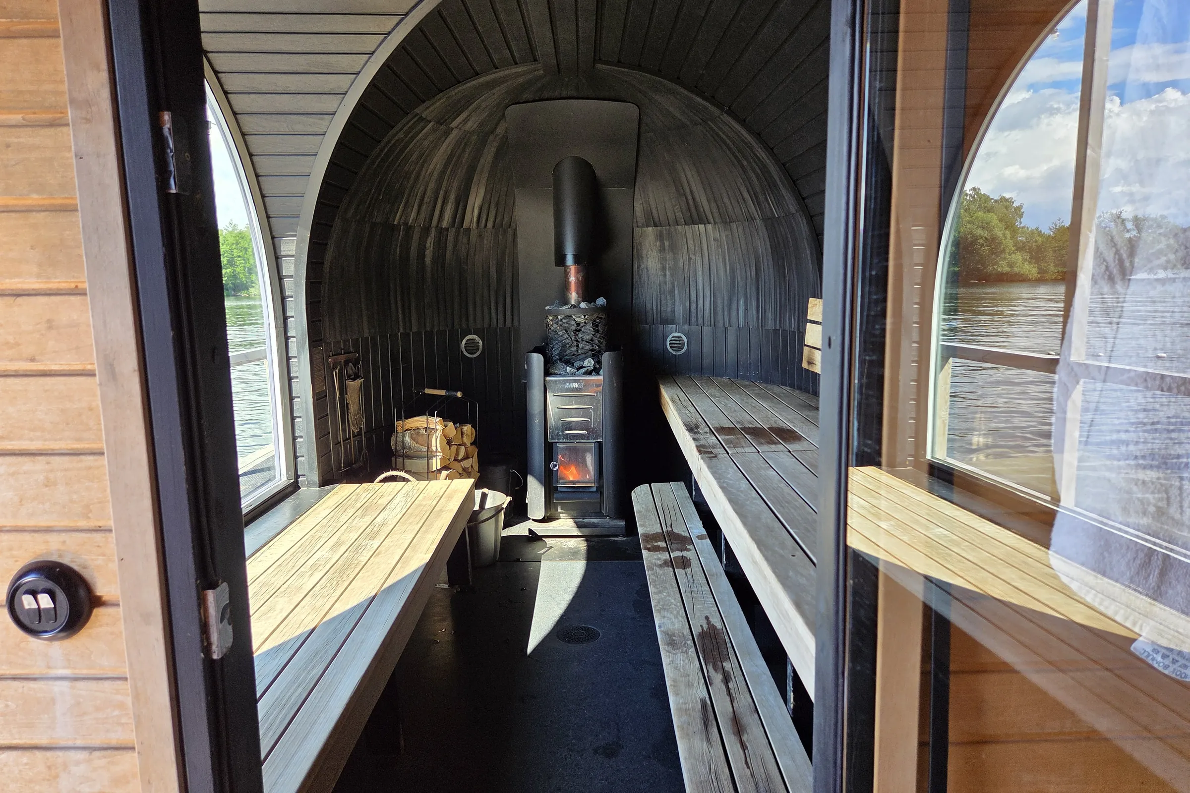 Sauna doorway and benches at STHLM Sauna Vinterviken View through the open door of the floating sauna, showing the arched black interior, benches on both sides, the wood-burning stove at the far end and arched windows looking out over the bay.