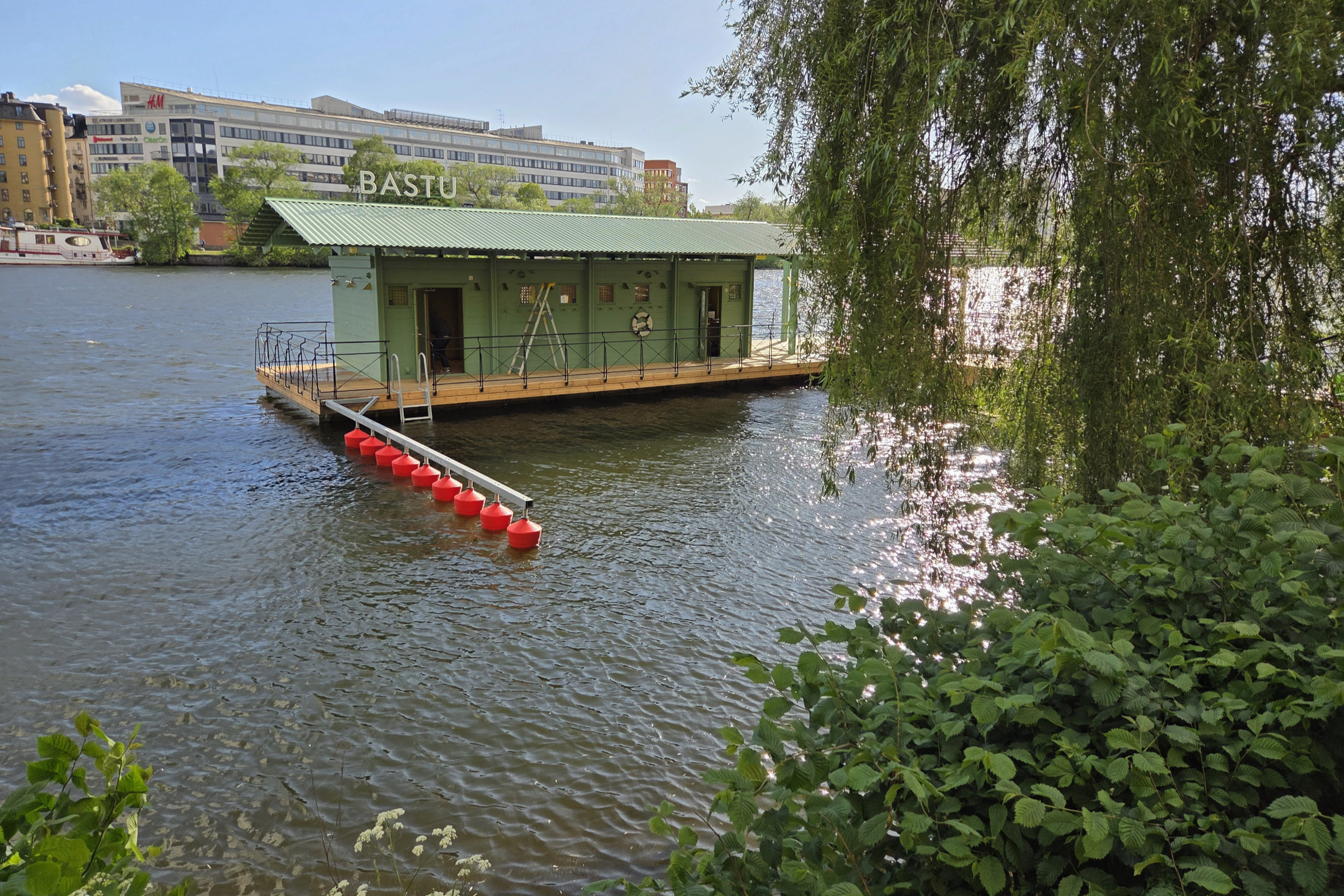 Hornstull Sauna in Stockholm floating on the water, with lush bushes and threes in the foreground.